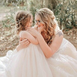 May include: A woman and a young girl embrace outdoors. Both wear floral crowns. The woman wears glasses and a white dress. The girl is in a light pink dress. The background is blurred with green foliage.