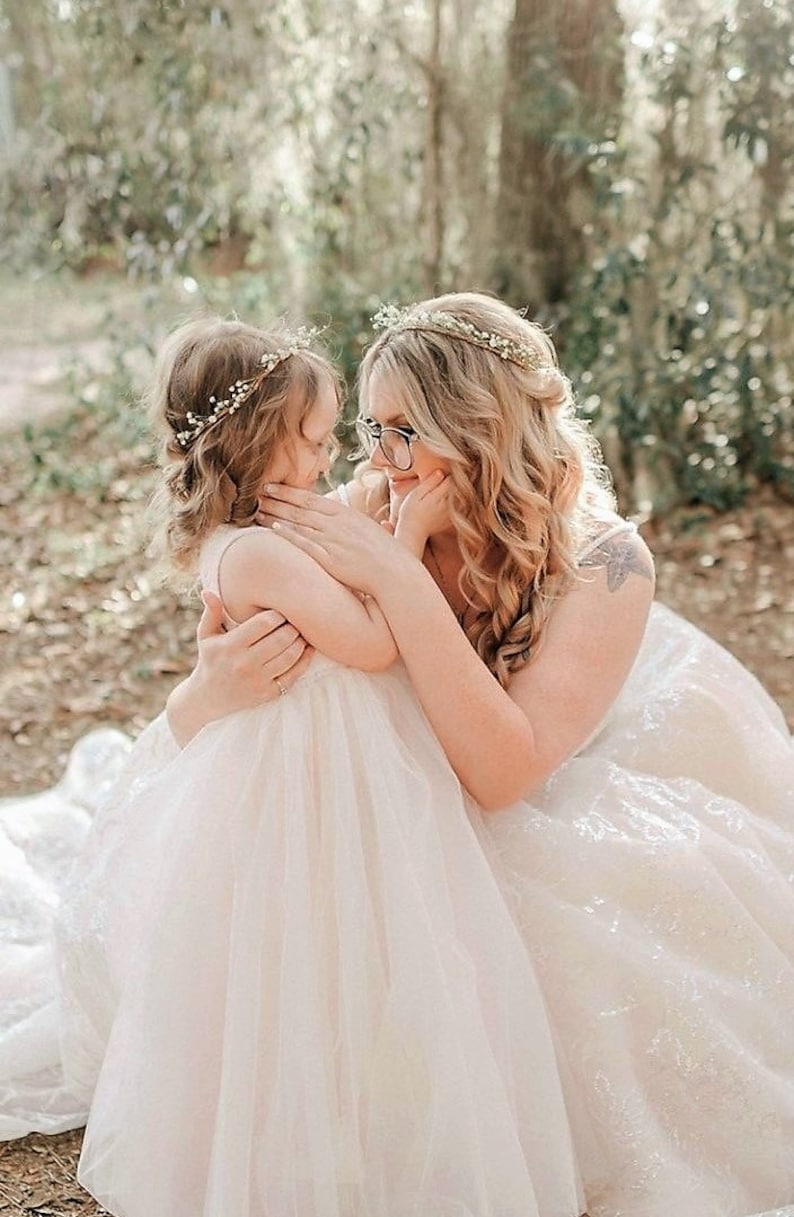 May include: A woman and a young girl embrace outdoors. Both wear floral crowns. The woman wears glasses and a white dress. The girl is in a light pink dress. The background is blurred with green foliage.