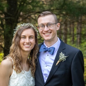 May include: A smiling couple poses outdoors in front of a blurred forest backdrop. The person on the left wears a white dress and floral crown. The person on the right wears a black suit, blue bow tie, and glasses.