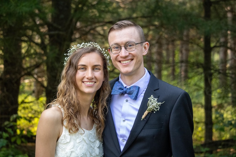 May include: A smiling couple poses outdoors in front of a blurred forest backdrop. The person on the left wears a white dress and floral crown. The person on the right wears a black suit, blue bow tie, and glasses.