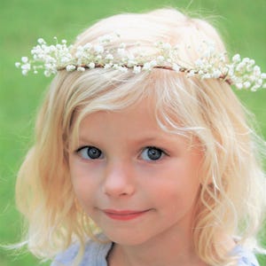 May include: A young person with blonde hair and blue eyes wearing a delicate floral crown made of small white flowers. The crown is set against the person's blonde hair, with a soft green background.