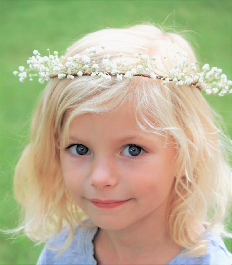 May include: A young person with blonde hair and blue eyes wearing a delicate floral crown made of small white flowers. The crown is set against the person's blonde hair, with a soft green background.