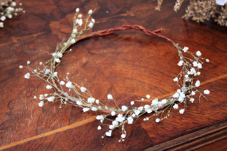 May include: A delicate floral crown featuring small white flowers and green stems, arranged on a brown, twisted wire base. The crown is set against a dark wooden surface, creating a natural and rustic aesthetic. Perfect for weddings or special occasions.
