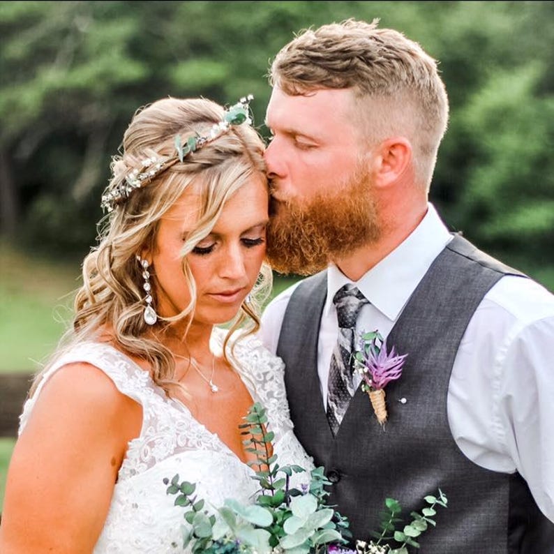 May include: A wedding portrait of a couple. The woman wears a white lace wedding dress, a floral headpiece, and dangling earrings. The man wears a gray vest, a patterned tie, and a boutonniere. The man is kissing the woman's forehead.