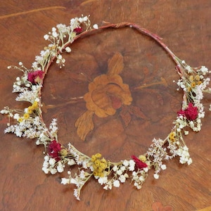 May include: A handcrafted floral crown featuring a woven base adorned with dried white, yellow, and burgundy flowers. The delicate arrangement is perfect for weddings, festivals, or bohemian-inspired events. The crown is displayed on a wooden surface.