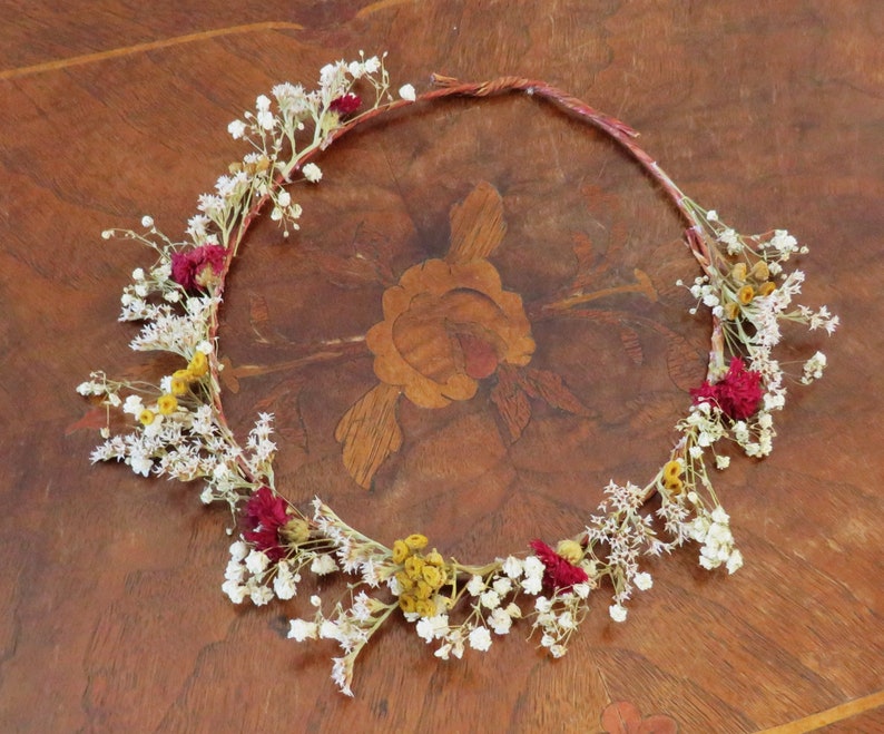 May include: A handcrafted floral crown featuring a woven base adorned with dried white, yellow, and burgundy flowers. The delicate arrangement is perfect for weddings, festivals, or bohemian-inspired events. The crown is displayed on a wooden surface.