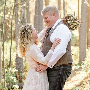 May include: A couple embraces in a sunlit forest. The woman wears a white lace dress and a floral crown. The man wears a white shirt, a tweed vest, and dark jeans. A bouquet of flowers rests on his back.