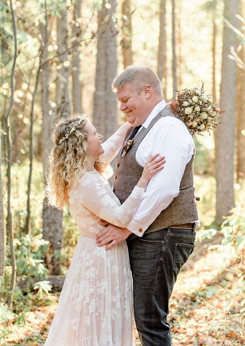 May include: A couple embraces in a sunlit forest. The woman wears a white lace dress and a floral crown. The man wears a white shirt, a tweed vest, and dark jeans. A bouquet of flowers rests on his back.