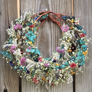 May include: A circular wreath composed of dried flowers in various colors, including white, purple, red, and teal. The wreath is made with a woven brown base and is displayed against a weathered wooden background. The wreath is a decorative item.