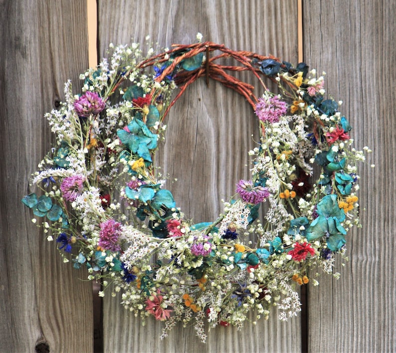 May include: A circular wreath composed of dried flowers in various colors, including white, purple, red, and teal. The wreath is made with a woven brown base and is displayed against a weathered wooden background. The wreath is a decorative item.