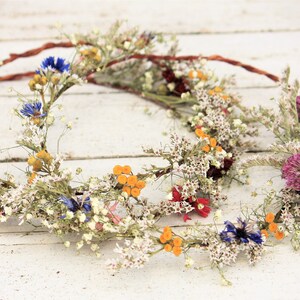 May include: Three floral crowns with dried flowers in various colors, including white, yellow, blue, and red. The crowns are arranged on a white wooden surface, showcasing their natural beauty and rustic charm.
