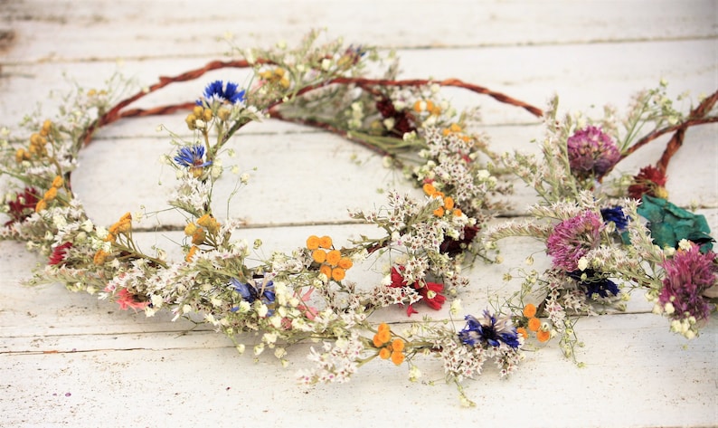 May include: Three floral crowns with dried flowers in various colors, including white, yellow, blue, and red. The crowns are arranged on a white wooden surface, showcasing their natural beauty and rustic charm.