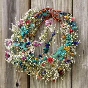 May include: A handmade floral wreath with a rustic aesthetic. The wreath features a variety of dried flowers in shades of red, pink, blue, yellow, and white, arranged on a brown vine base. The wreath is displayed against a weathered wooden background.