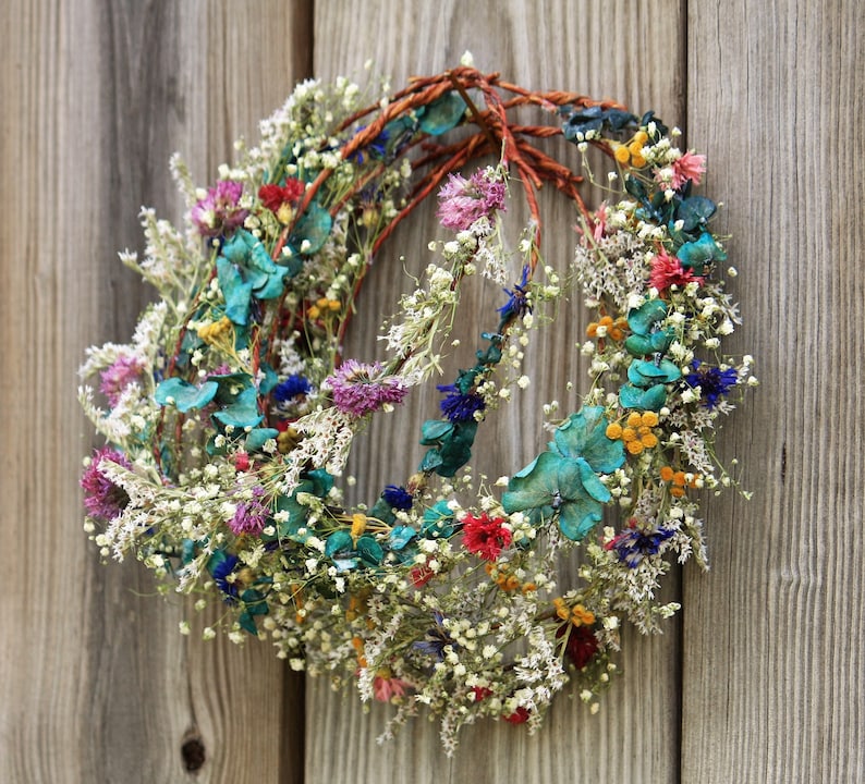 May include: A handmade floral wreath with a rustic aesthetic. The wreath features a variety of dried flowers in shades of red, pink, blue, yellow, and white, arranged on a brown vine base. The wreath is displayed against a weathered wooden background.