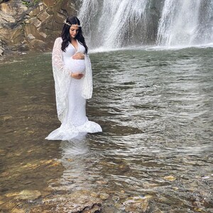 May include: A white lace maternity gown with long sleeves and a plunging neckline. The dress is worn by a person standing in a stream of water in front of a waterfall. A floral crown adorns the person's head.