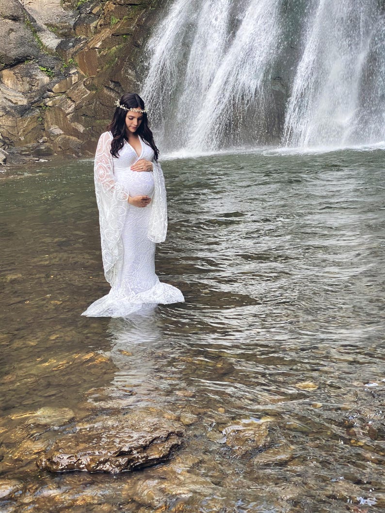 May include: A white lace maternity gown with long sleeves and a plunging neckline. The dress is worn by a person standing in a stream of water in front of a waterfall. A floral crown adorns the person's head.
