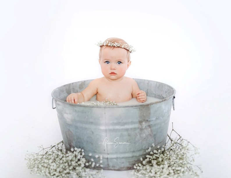 May include: A baby sits in a galvanized metal tub filled with milky water, wearing a floral crown. White flowers are scattered around the tub. The baby has fair skin and blue eyes. The image is set against a white backdrop.