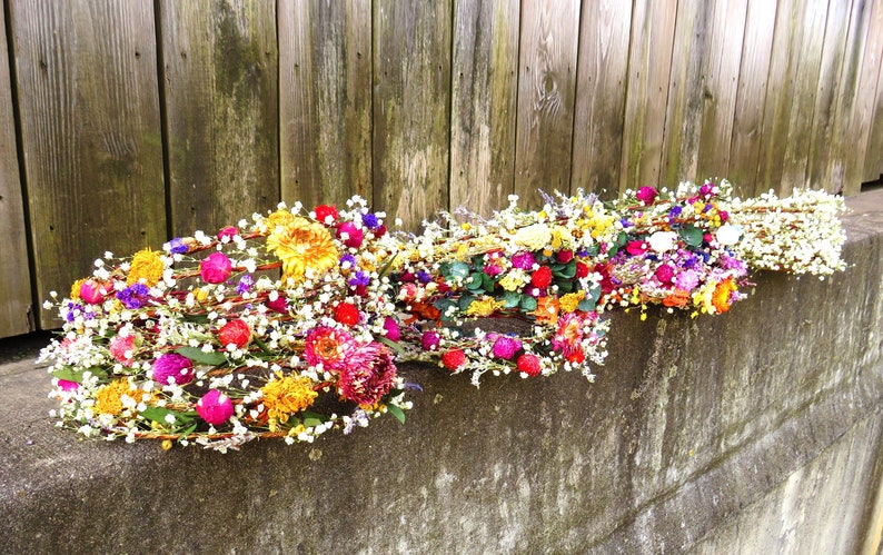 May include: Several colorful floral crowns rest on a concrete ledge in front of a wooden fence. The crowns feature a variety of flowers in shades of yellow, pink, red, purple, and white. The crowns are made of woven branches.