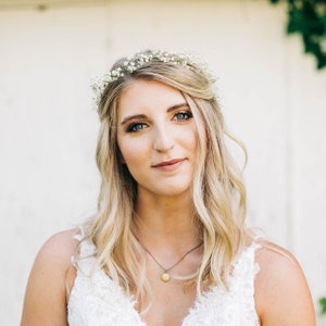 May include: A woman with blonde hair and blue eyes wearing a delicate flower crown. She is wearing a white lace dress and a gold pendant necklace. The background is a blurred white wall.