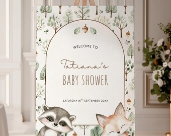 May include: A baby shower welcome sign on an easel. The sign features a woodland theme with a raccoon and fox illustration. The text reads "Welcome to Tatiana's Baby Shower" with the date. The background has a pattern of trees and acorns.