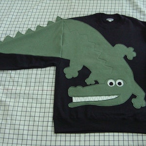 May include: A black sweatshirt with a green alligator applique on the front. The alligator has a large mouth with white teeth and two large white eyes. The alligator's tail extends across the left shoulder of the sweatshirt.