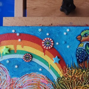 May include: A colorful painting featuring a rainbow, a bird, and decorative elements. The bird has green, orange, and blue feathers. The artwork includes star and circular embellishments against a blue background.