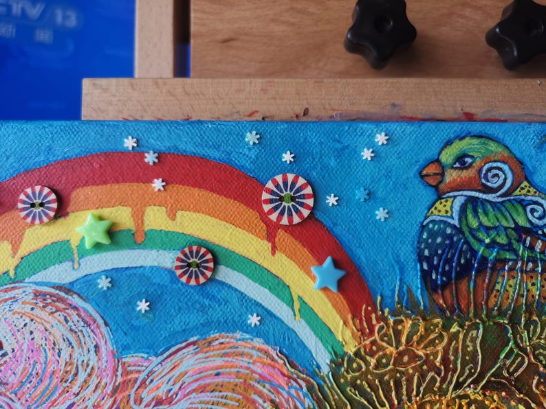 May include: A colorful painting featuring a rainbow, a bird, and decorative elements. The bird has green, orange, and blue feathers. The artwork includes star and circular embellishments against a blue background.