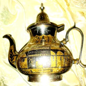 May include: A vintage metal teapot with a decorative design in gold and black. The teapot has a curved spout, a handle, and a lid with a small finial. The body of the teapot features an etched pattern. The teapot is likely made of brass or a similar metal.