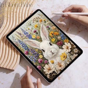 May include: A digital art print on a tablet featuring a white rabbit surrounded by colorful wildflowers. The artwork includes yellow, purple, and white flowers. The tablet is held by a person's hand, with the text "In Bloom Babe" in the upper left corner.