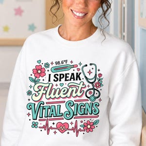 May include: A white long-sleeve sweatshirt featuring a colorful graphic print that reads I SPEAK Fluent VITAL SIGNS. The design includes a thermometer showing 98.6°F, a stethoscope, a heart with an EKG line, and various small pink and teal flowers.