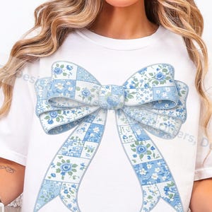 Puede incluir: Camiseta blanca con un gran diseño de lazo patchwork azul claro. El lazo tiene un estampado floral dentro de los cuadrados de patchwork. El texto en la parte inferior dice "Patchwork Coquette T-Shirt PNG Design". La camiseta es de estilo casual.