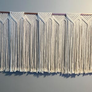May include: Cream-colored macrame wall hanging. The design features a series of triangular patterns across the top, with long, vertical fringe hanging below. The macrame is suspended from a wooden dowel with copper accents, creating a bohemian aesthetic.