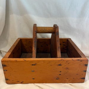 May include: A vintage wooden carrier with a handle and three compartments. The carrier is made of light brown wood with visible nail heads and signs of wear, suggesting age and use. The handle is centered, allowing for easy transport.