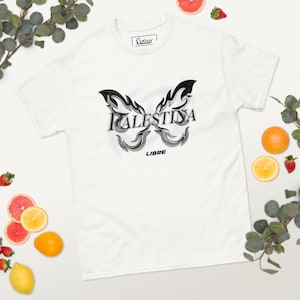 Puede incluir: Camiseta blanca con un diseño de mariposa. La palabra "Palestina" está en el centro del diseño de la mariposa, con la palabra "LIBRE" debajo. El diseño de la mariposa es negro y gris.
