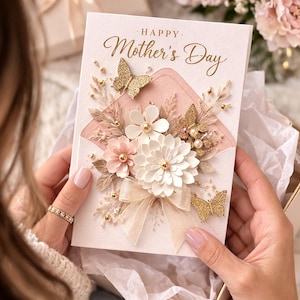 May include: A Mother's Day card with a floral design and gold glitter butterflies. The card features the words "Happy Mother's Day" in elegant script. The card is held in a gift box with a bow.