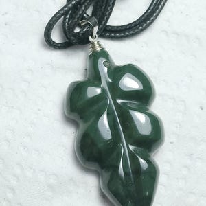 May include: A dark green leaf-shaped pendant necklace. The pendant is made of a polished, possibly jade, material and is suspended from a black cord necklace. The leaf design has detailed veining and a smooth, glossy finish.