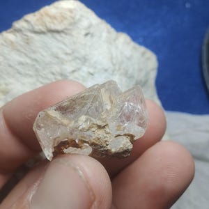 Puede incluir: Un espécimen mineral claro y cristalino con facetas geométricas afiladas. La piedra se sostiene sobre un fondo neutro, mostrando su transparencia y estructura interna. El mineral parece ser un cristal de cuarzo, con una base áspera y terrosa.