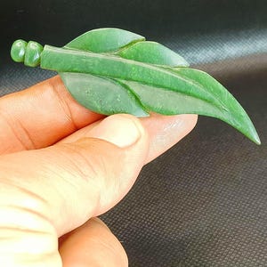 Könnte beinhalten: Ein geschnitzter Jade-Blatt-Anhänger in einem leuchtenden Grünton. Das Blattdesign weist detaillierte Adern und einen Stiel mit abgerundetem Ende auf. Der Anhänger wird in einer Hand vor dunklem Hintergrund gehalten.
