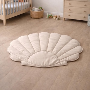 Könnte beinhalten: Cremefarbener, muschelförmiger Teppich mit Steppmuster auf hellem Holzboden. Das Zimmer enthält ein Kinderbett, eine Kommode und Spielzeug, was auf ein Kinderzimmer hindeutet. Der Teppich hat einen Durchmesser von ca. 120 cm.