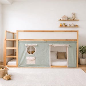 May include: A wooden loft bed with a light green fabric curtain, a round window, and a pocket. The bed has a ladder and a lower bed area. The room includes a rug, shelves with toys, and a small table.