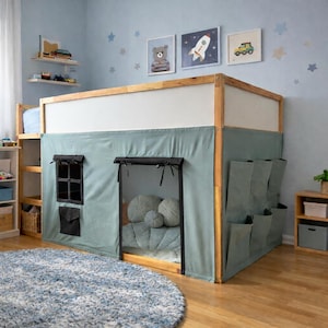 May include: A wooden loft bed with a light blue tent-like enclosure. The bed has a window and door opening, with pockets on the side. The room has a blue rug, star decals, and framed artwork.