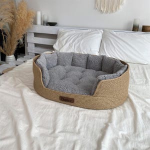 May include: Oval-shaped dog bed with a woven, light brown exterior and a soft, gray interior. The bed has a low profile and a rectangular label that reads "Buster". The bed is placed on a white sheet.