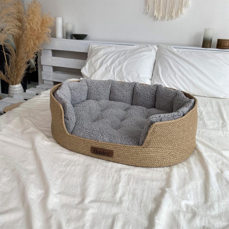 May include: Oval-shaped dog bed with a woven, light brown exterior and a soft, gray interior. The bed has a low profile and a rectangular label that reads "Buster". The bed is placed on a white sheet.