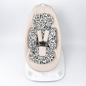 Puede incluir: Un balancín para bebé beige con reposacabezas y asiento con estampado de leopardo. El balancín tiene una base blanca con botones de control y altavoces. El asiento tiene un arnés de cinco puntos.