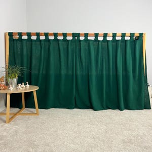 May include: Dark green curtains hanging from a wooden frame. The curtains are made of a textured fabric and are gathered at the top. A small round table with a plant and figurines is in front of the curtains.