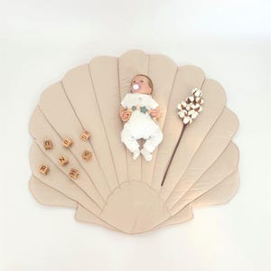 Op de afbeelding: Een baby ligt op een beige schelpvormige speelmat. De baby draagt een witte onesie en heeft een fopspeen. Houten letterblokken en een katoenen stengel liggen ook op de mat. De mat ligt op een witte achtergrond.