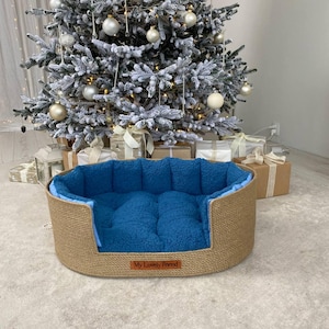 May include: Oval pet bed with a woven jute exterior and a plush blue interior. The bed has a low profile and a rectangular shape. The words "My Lovely Friend" are on a small brown tag on the front of the bed. A Christmas tree is in the background.
