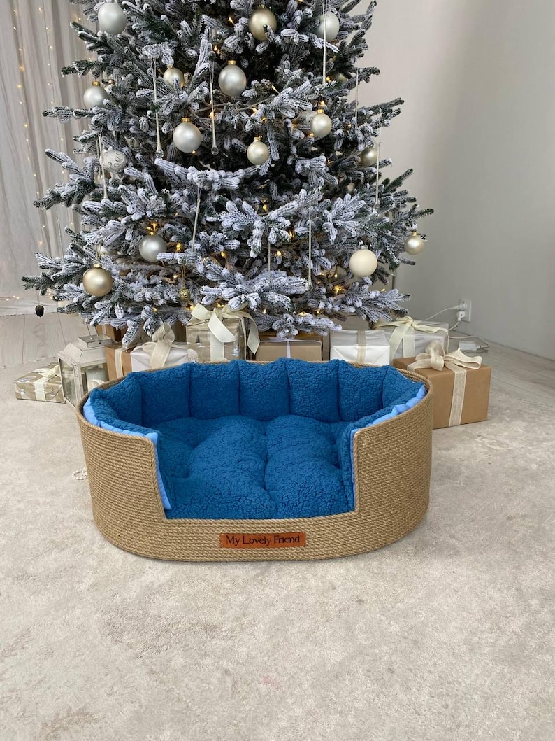 May include: Oval pet bed with a woven jute exterior and a plush blue interior. The bed has a low profile and a rectangular shape. The words "My Lovely Friend" are on a small brown tag on the front of the bed. A Christmas tree is in the background.
