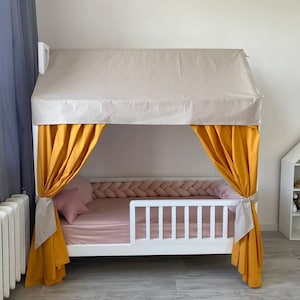 May include: A white wooden house-shaped bed frame featuring a beige fabric roof cover, mustard yellow curtains tied to the sides, a pink fitted sheet, a pink star-shaped pillow, and a braided pink fabric bumper along the interior frame.