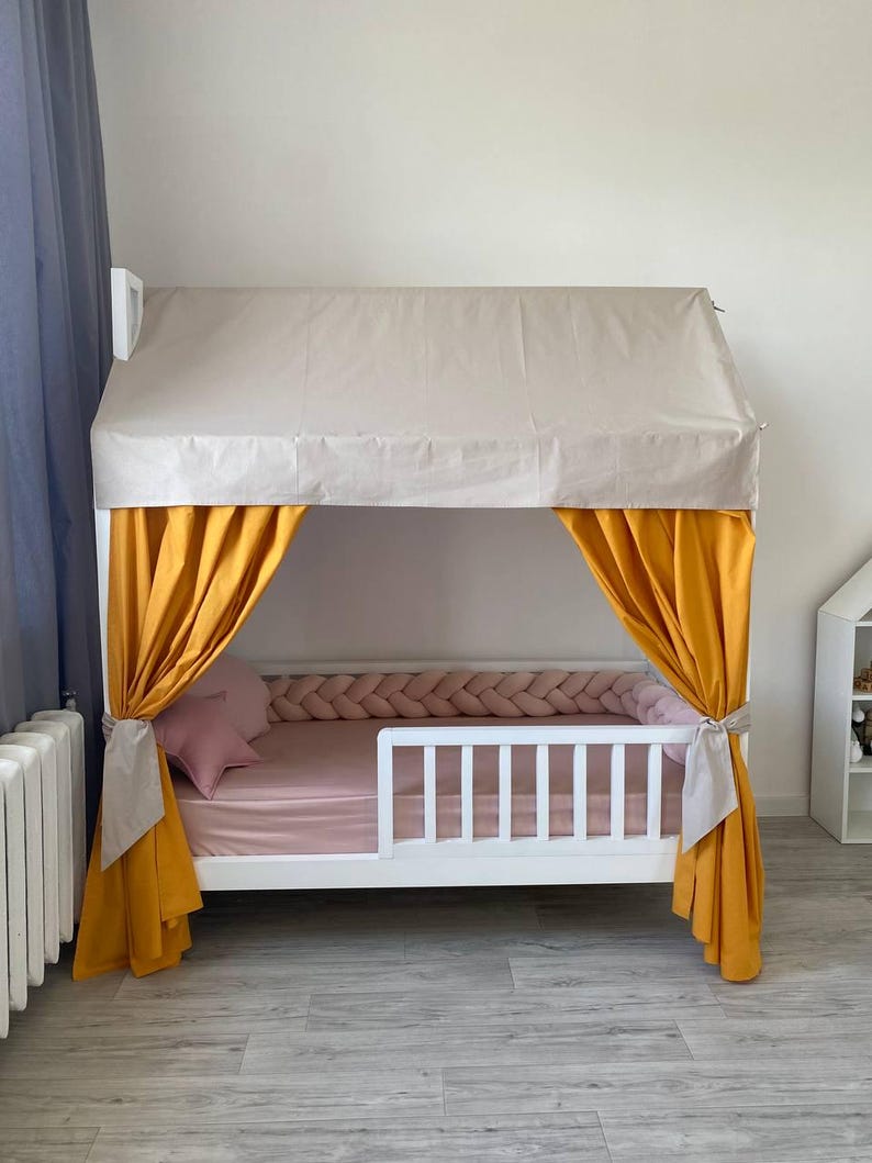 May include: A white wooden house-shaped bed frame featuring a beige fabric roof cover, mustard yellow curtains tied to the sides, a pink fitted sheet, a pink star-shaped pillow, and a braided pink fabric bumper along the interior frame.
